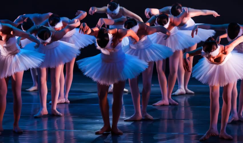 A large group of ballet dancers perform on a dark stage