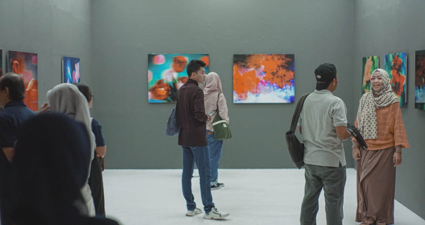 A crowd of art lovers stand and chat in a gallery filled with bright paintings