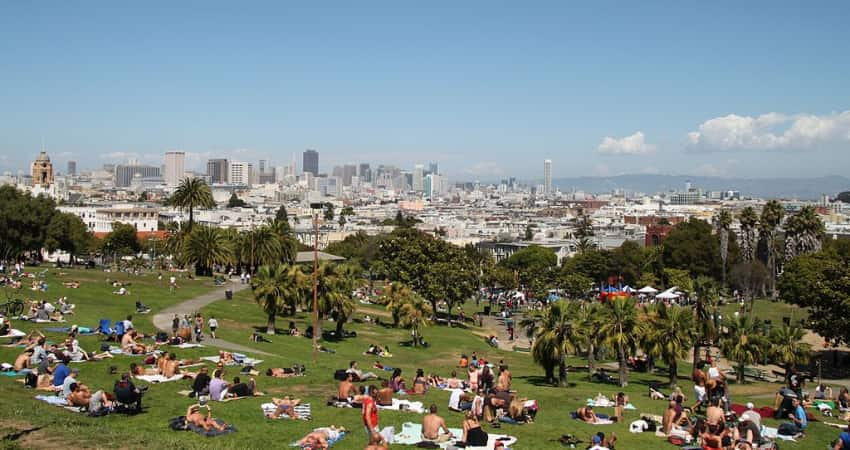 Dozens of people sunbathing on a hill in Dolores Park with the San Francisco skyline in the background