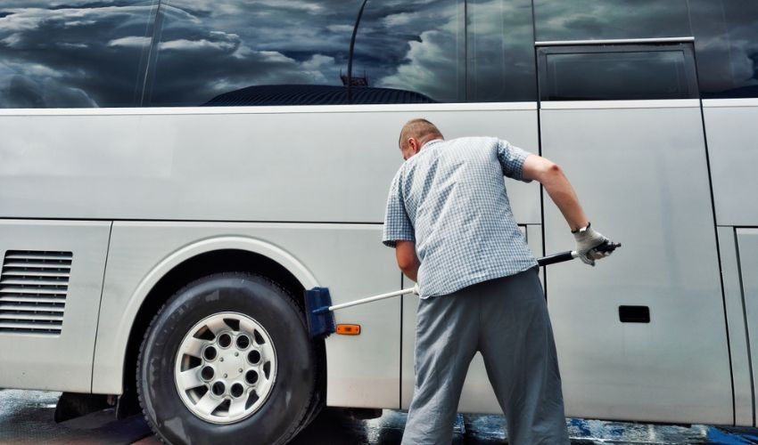 Charter bus operator cleaning the wheel well of a charter bus with a mop.